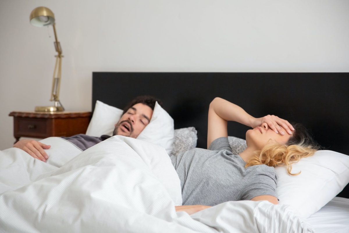 Woman holding head as man snores beside her in bed
